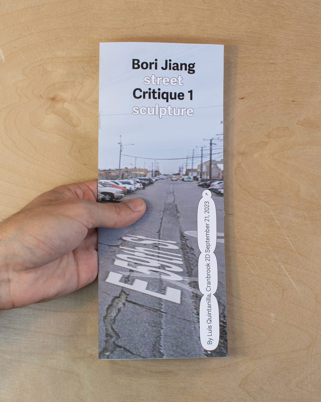 hands holding a zine of a critical review of Bori Jiang, cover features an image of a street in los angeles with the title centered towards the top in black and white over a wood panel background